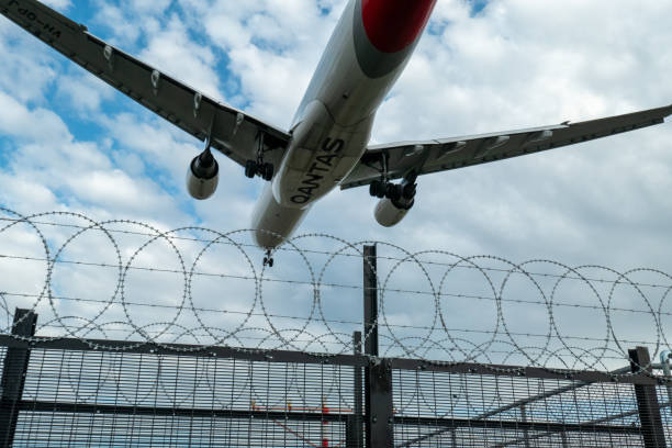 A Qantas airplane flies low over a barbed wire fence during landing, with a partly cloudy sky in the background.