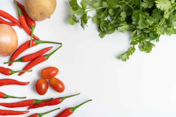 Layflat composition featuring vibrant red chili peppers, onion, potato, cherry tomatoes and corianders with a copy space against a clean, white backdrop.