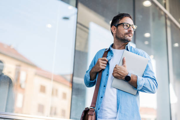 istock A handsome, cheerful, smiling man walking by a building, carrying a laptop. 2170783620