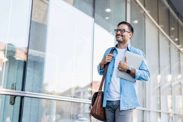 istock A handsome, cheerful, smiling man walking by a building, carrying a laptop. 2170783682