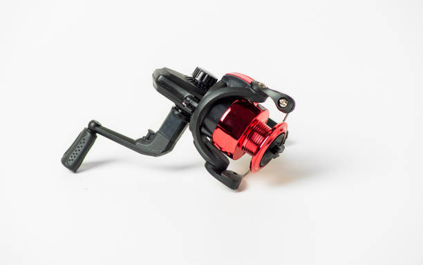 A red and black fishing reel sits on a white background. The reel is small and has a black handle