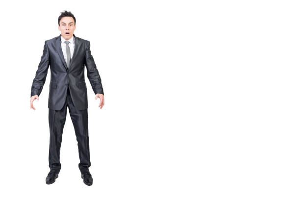 Full length of shocked young male manager in formal suit and tie with dark hair looking at camera with opened mouth while standing in studio against white background