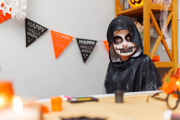 Child wearing black robe and skeleton makeup at a Halloween party, sitting near a decorated table with Halloween banners and decorations in the background. Indoor portrait