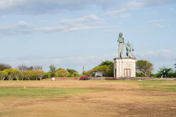A large statue of two figures stands on a grassy area with sparse vegetation. The sky is partly cloudy, and there are some trees and shrubs in the background. The ground appears dry and brown, indicating a lack of water.