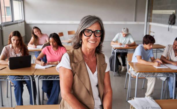 istock Portrait of a mature teacher at work looks at camera smiling while students study in the background. 2170790345