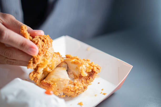 A close-up of hands holding a piece of crispy fried chicken, showcasing the golden-brown coating and juicy meat inside. The background is blurred, emphasizing the food.