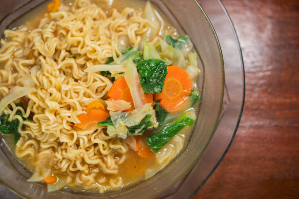 A bowl of ramen noodles with vegetables including carrots and spinach, served on a wooden table. In the background, another plate of food is slightly out of focus.