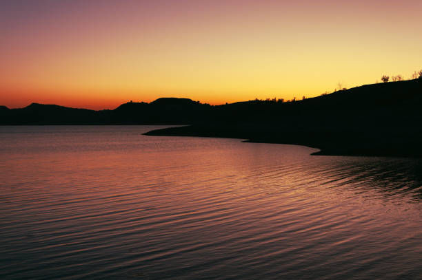 sun sets, painting sky and reflecting on serene lake with hills in background, creating calming atmosphere with vibrant colors