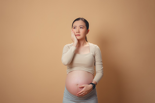 Asian Pregnant Woman Thinking About Problem Worried And Confused Isolated On Beige Background Stock Photo - Download Image Now - iStock