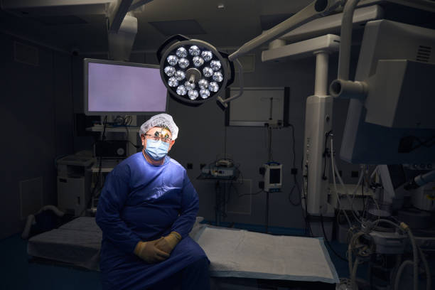 Man in blue surgical scrubs stands in an empty operating room lit by overhead operating lights