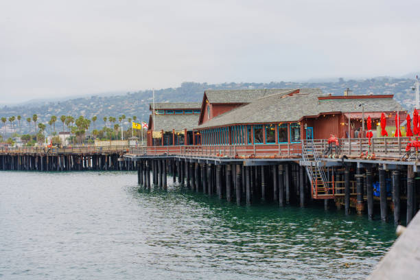 Santa Barbara, California - April 20, 2024: Scenic view of Stearns Wharf featuring waterfront dining, colorful flags, and palm trees along the calm waters of Santa Barbara