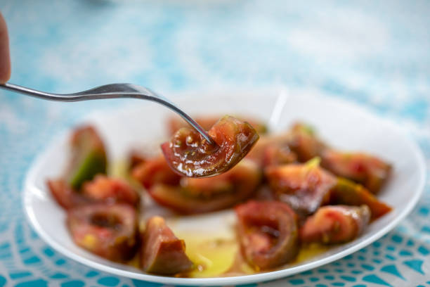 Plate of split tomato for salad on table with blue tablecloth and a hand with fork in daylight