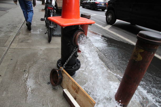 A fire hydrant bursts forth with water on a bustling New York City street as pedestrians walk past. Traffic continues amid the spray, highlighting urban life.