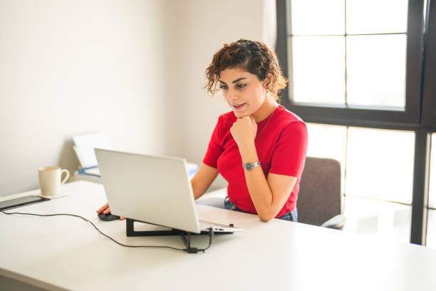 curly haired woman in red t-shirt sitting at the computer desk taking notes while looking at her laptop