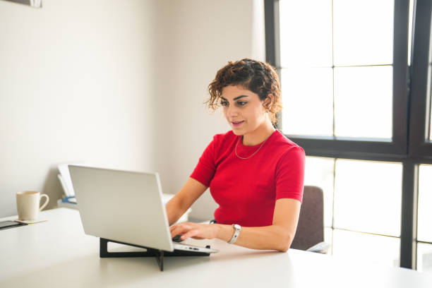 istock curly haired woman in red t-shirt sitting at the computer desk taking notes while looking at her laptop 2170850425