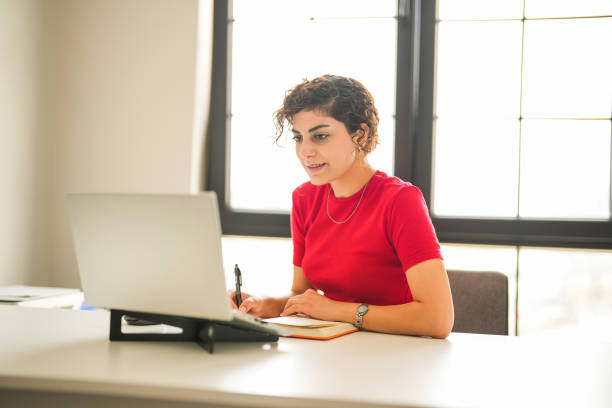 istock curly haired woman in red t-shirt sitting at the computer desk taking notes while looking at her laptop 2170851159