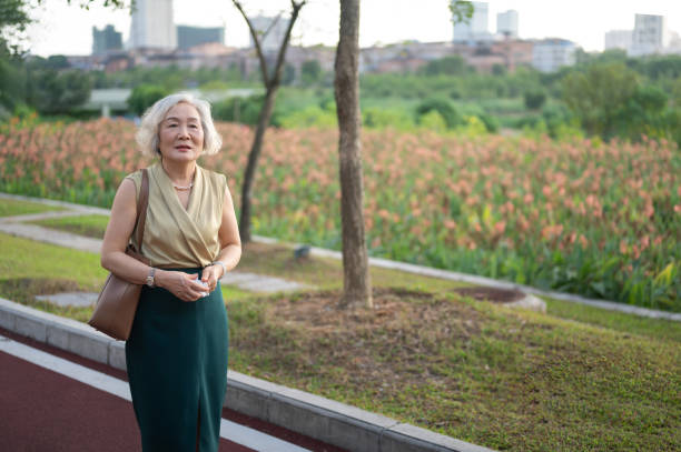Senior Asian woman with gray hair stands in a park with blooming flowers, holding her purse, and a cityscape forms the backdrop, demonstrating a mix of nature and urban life around her.