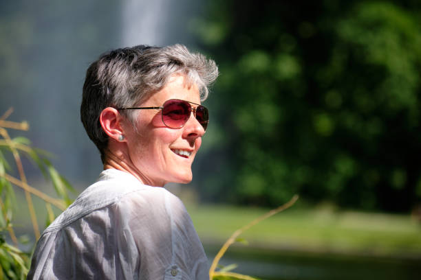 A smiling woman with short gray hair and sunglasses enjoys a sunny day outdoors, looking relaxed and content. The natural setting, with blurred greenery and sunlight reflecting off her white shirt, conveys a peaceful and carefree atmosphere. Her smile and relaxed posture highlight the simple pleasure of being in the moment.