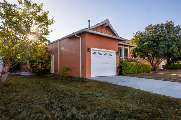 Charming suburban home, perfect for real estate, lifestyle, or interior design projects. The facade features wood siding painted in a warm cream hue, complemented by a brown tiled roof and white chimneys. The front garden, with manicured green grass and a flowering tree with delicate pink blossoms, adds a serene, spring-like touch.