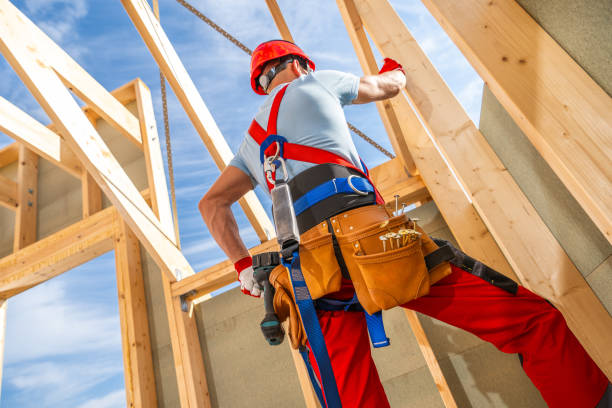 A construction worker in a red hard hat and tool belt is busy securing wooden beams in a partially built structure under a clear blue sky.