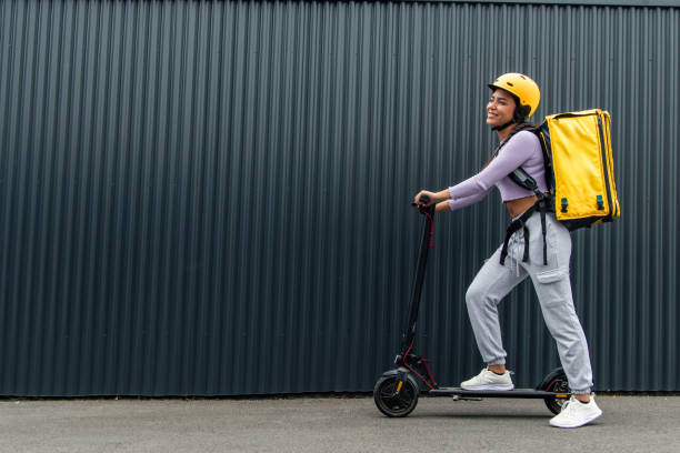 istock copyspace Smiling Latin courier with yellow backpack on electric scooter in front of black fence 2170876630