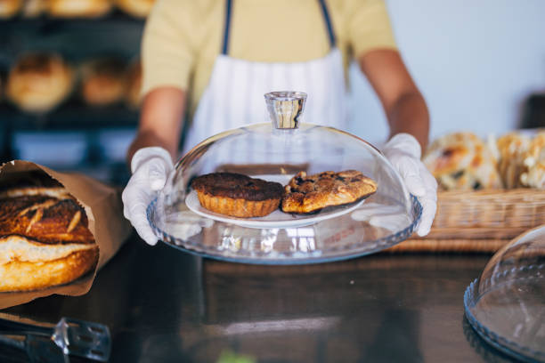 A baker wearing gloves displays two delicious pastries under a glass dome, showcasing freshness in a cozy bakery setting.