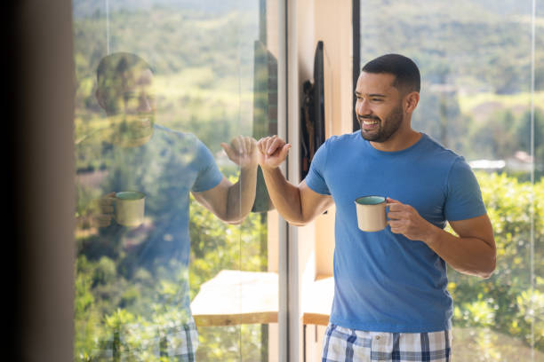 Happy Latin American man at home drinking a cup of coffee while looking through the window and smiling