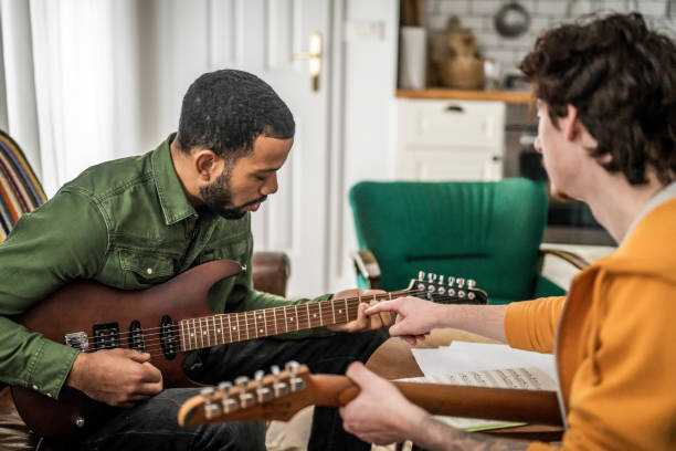 A Caucasian instructor guides a Black student in a guitar playing session, both focused on the instrument, in a cozy indoor setting.
