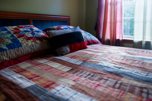 Welcoming bedroom features a comfortable bed with colorful patchwork quilts in red, blue, and neutral tones. Soft natural light filters through sheer curtains, highlighting the wooden headboard and subtle wall color. This cozy space embodies the rustic charm of a middle-class Poconos retreat.