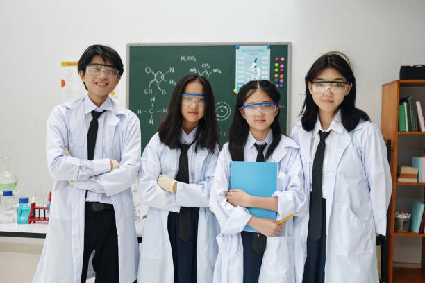 Asian students standing in science classroom wearing lab coats and safety glasses while holding science materials. They are surrounded by lab equipment and a chalkboard