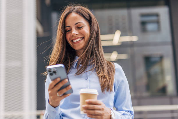 istock A young, cheerful businesswoman, smiling while holding coffee  and using her smartphone on the city street, with modern buildings in the background. 2170929521