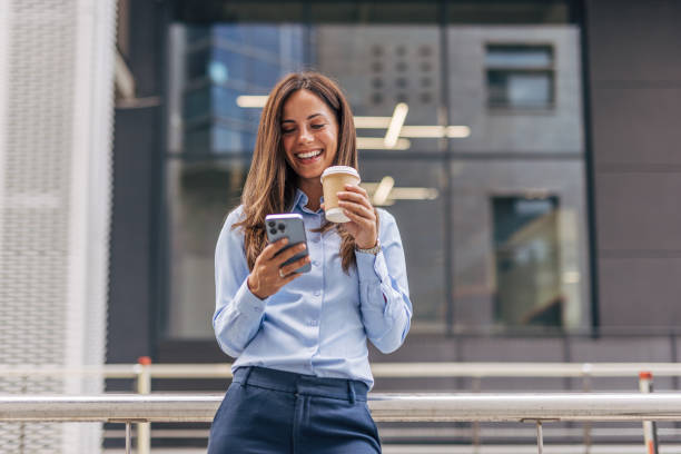 istock A young, cheerful businesswoman, smiling while holding coffee  and using her smartphone on the city street, with modern buildings in the background. 2170929588