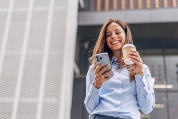istock A young, cheerful businesswoman, smiling while holding coffee  and using her smartphone on the city street, with modern buildings in the background. 2170929606