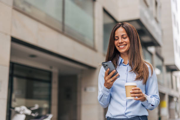istock A young, cheerful businesswoman, smiling while holding coffee  and using her smartphone on the city street, with modern buildings in the background. 2170930159