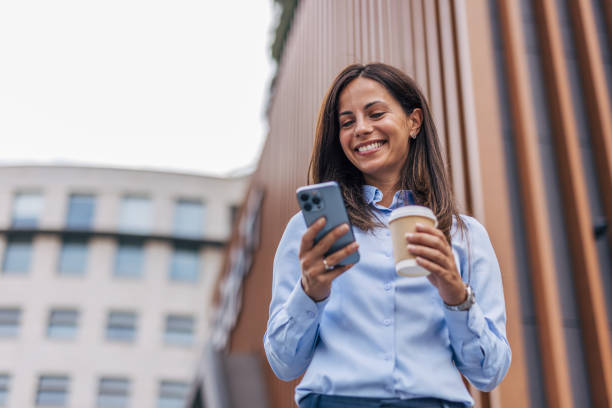 istock A young, cheerful businesswoman, smiling while holding coffee  and using her smartphone on the city street, with modern buildings in the background. 2170930423