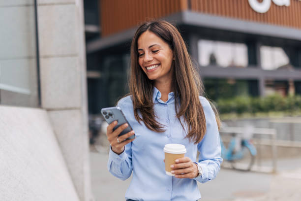 istock A young, cheerful businesswoman, smiling while holding coffee  and using her smartphone on the city street, with modern buildings in the background. 2170930544