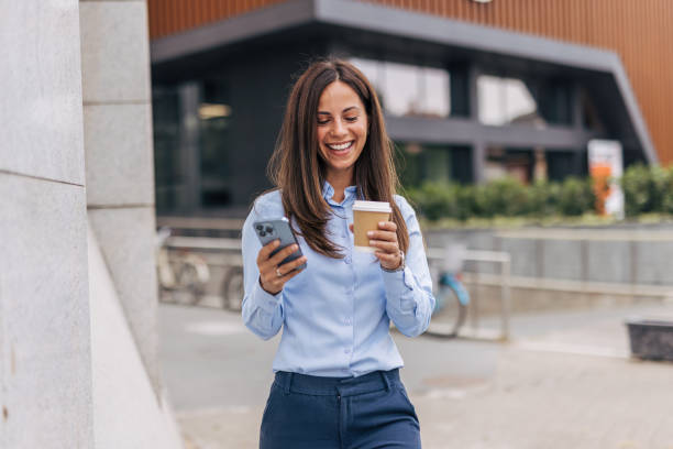 istock A young, cheerful businesswoman, smiling while holding coffee  and using her smartphone on the city street, with modern buildings in the background. 2170930569
