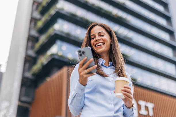 istock A young, cheerful businesswoman, smiling while holding coffee  and using her smartphone on the city street, with modern buildings in the background. 2170930794
