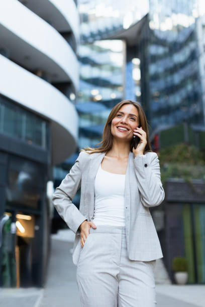 istock Woman in a light gray suit happily talking on her phone while walking past contemporary architecture in a bustling urban area 2170931921