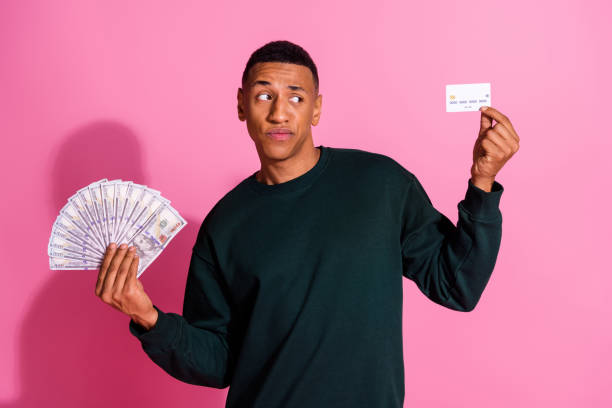 istock Photo of dountful uncertain guy dressed pullover comparing bank card dollars fan emtpy space isolated pink color background 2170934550