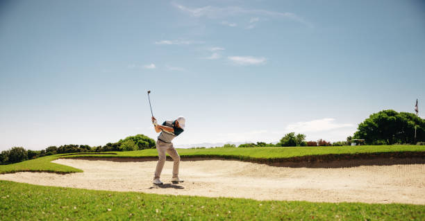 Male golfer swinging golf club in bunker escape during match A male golfer skillfully swinging a golf club to escape a bunker during a competitive match, capturing determination and precision. game swing stock pictures, royalty-free photos & images