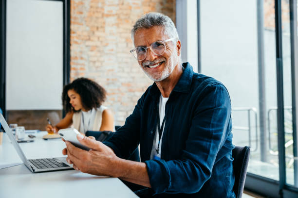 Seasoned business executive confidently working in a modern office setting, smiling while using a smartphone and laptop