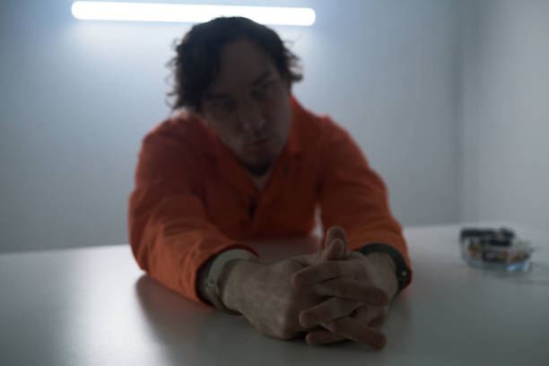 Man sitting at table, hands clasped, wearing orange jacket, with dim lighting highlighting his features and casting shadows in room