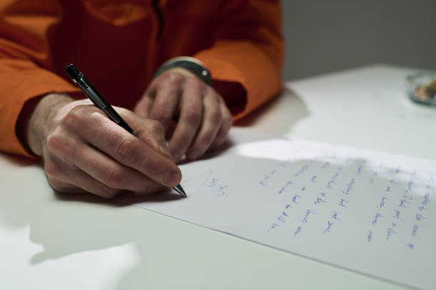 Hand holding pen and writing on paper, featuring content written in cursive script on sheet placed on light surface, with wristwatch visible