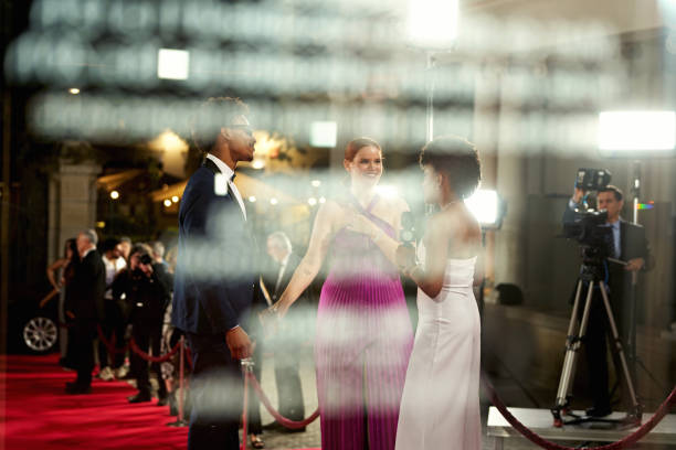 Personal perspective of red carpet correspondent holding microphone and standing with smiling couple, conversing, everyone in evening wear.
