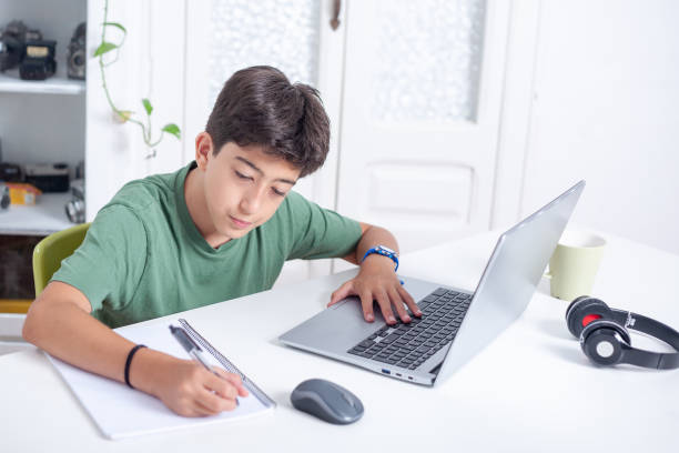istock A young boy is sitting at a desk with a laptop computer and writing in a notebook with a pen. 2170988091