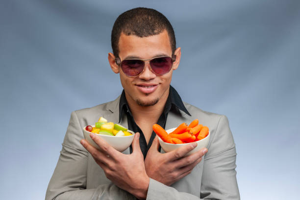 Head and shoulders studio shot of a smiling cool man with sunglasses holding a bowl with carrots and a bowl with fresh fruits on gray background