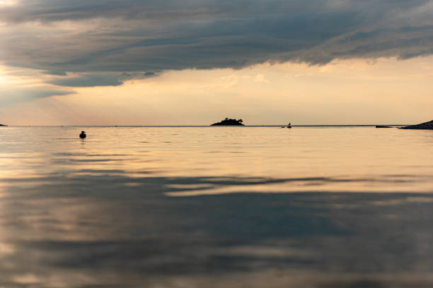 Serene view of a lone sailboat drifting on calm waters under a vibrant sunset sky. The horizon glows with warm orange and pink tones, framed by distant landmasses and a peaceful atmosphere. Ideal for visuals focused on solitude, natural beauty, maritime lifestyle, and scenic ocean moments. No specific location indicated.