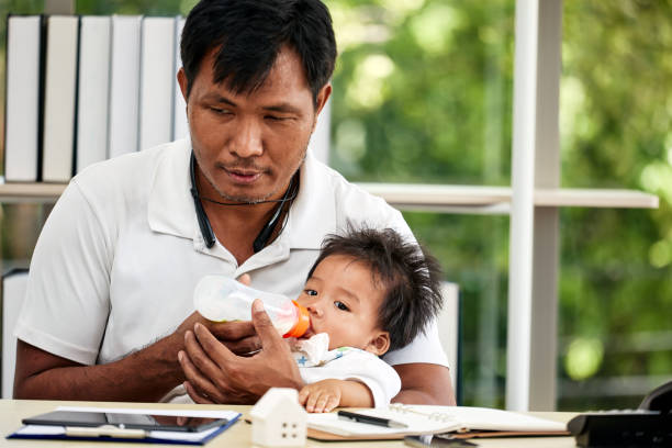 istock Asian father feeding baby with bottle holding securely on his lap while sitting at desk. home office, with notebooks, tablet, and miniature house model on table. calm drinking from bottle 2171005977