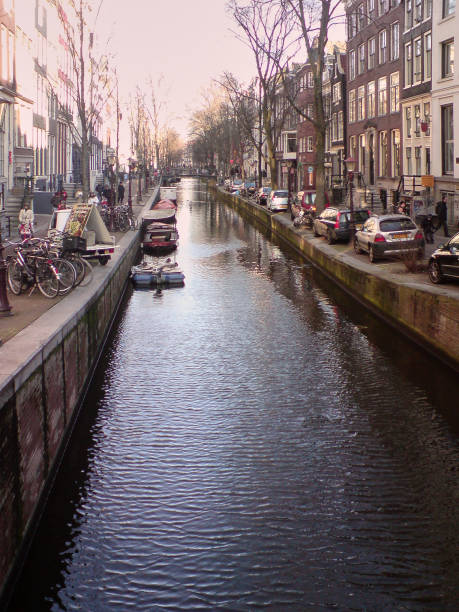 This image shows a picturesque canal scene typical of cities like Amsterdam, with calm waters running between rows of historic buildings on either side. Along the canal, boats are moored, and bicycles are parked along the edge, emphasizing the city's bike-friendly culture. The street is lined with cars and narrow sidewalks, where pedestrians can be seen enjoying a walk. The architecture reflects traditional Dutch canal houses, tall and narrow with large windows and brick facades. The lighting suggests that it may be late afternoon, casting a soft glow over the scene, enhancing its peaceful, everyday charm.